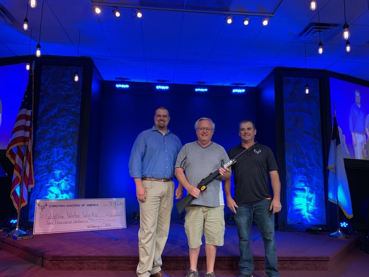 Three Christian men standing on stage with a rifle.